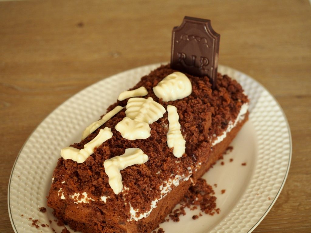 Ein Mailwurfkuchen in Form eines Grabes mit einem Grabstein aus dunkler Schokolade und einem Skelett aus weißer Schokolade