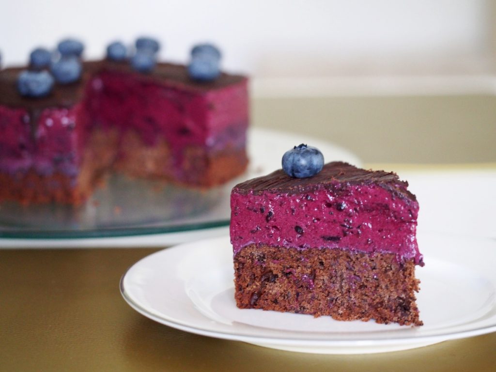 Ein Stück einer Blaubeer-Torte auf einem Kuchenteller im Vordergrund. Dahinter die angeschnittene Torte auf einem Tortenteller