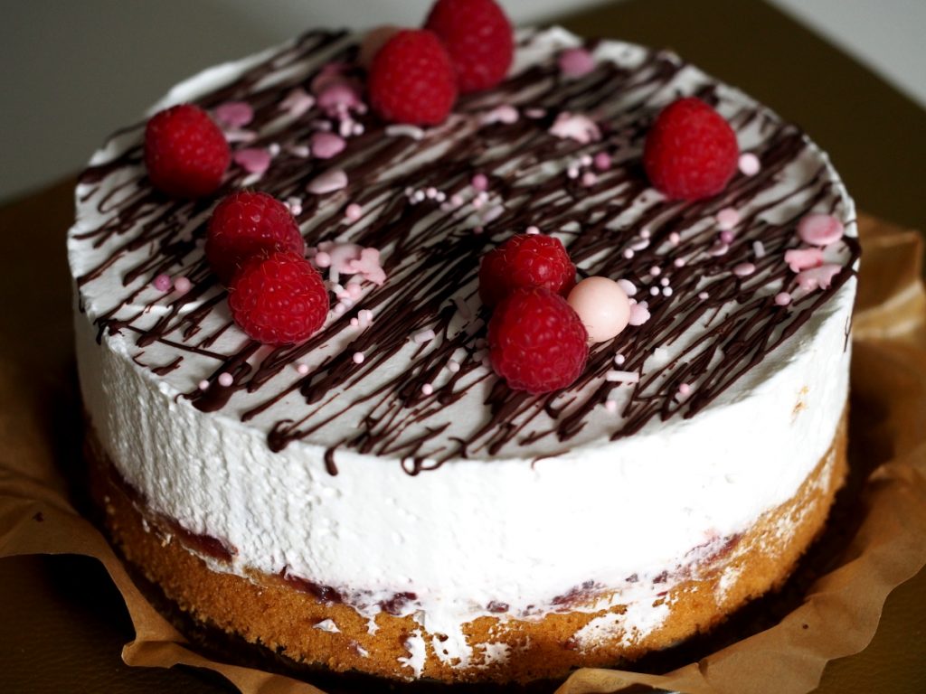Die Himbeertorte im Ganzen, dekoriert mit Schokoladensprenkel, Zuckerstreusel in den Farben rosa und Weiß und frischen Himbeeren, die ungleichmäßig auf der Oberfläche verteilt wurden