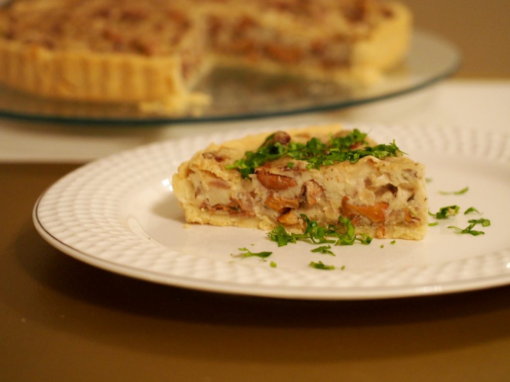 Ein Stück der Pfifferling Quiche auf einem Teller, garniert mit frischer Petersilie. Im Hintergrund die angeschnittene Quiche