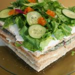 Die quadratische Sandwichtorte auf einer gläsernen Tortenplatte. Die Sandwichtorte wurde mit Salat, Gurke und veganem Kaviar dekoriert.