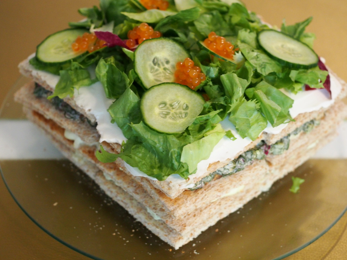 Die quadratische Sandwichtorte auf einer gläsernen Tortenplatte. Die Sandwichtorte wurde mit Salat, Gurke und veganem Kaviar dekoriert.