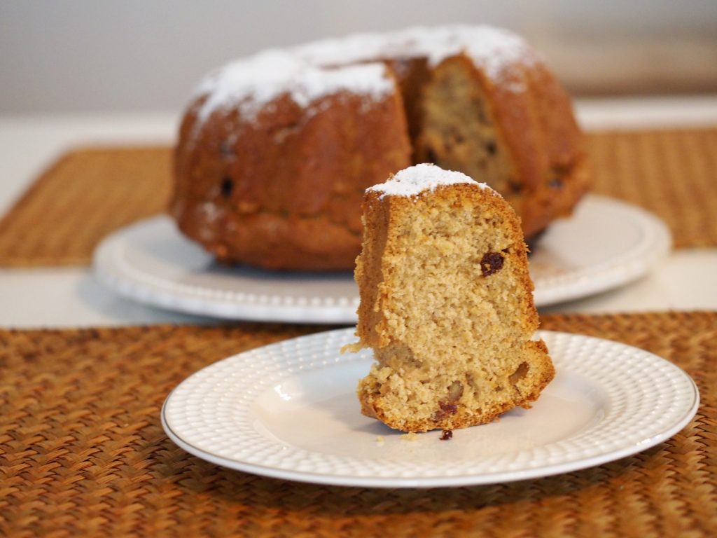 Ein Stück des Gugelhupf im Vordergrund auf einem kleinen Teller, dahinter der angeschnittene Kuchen