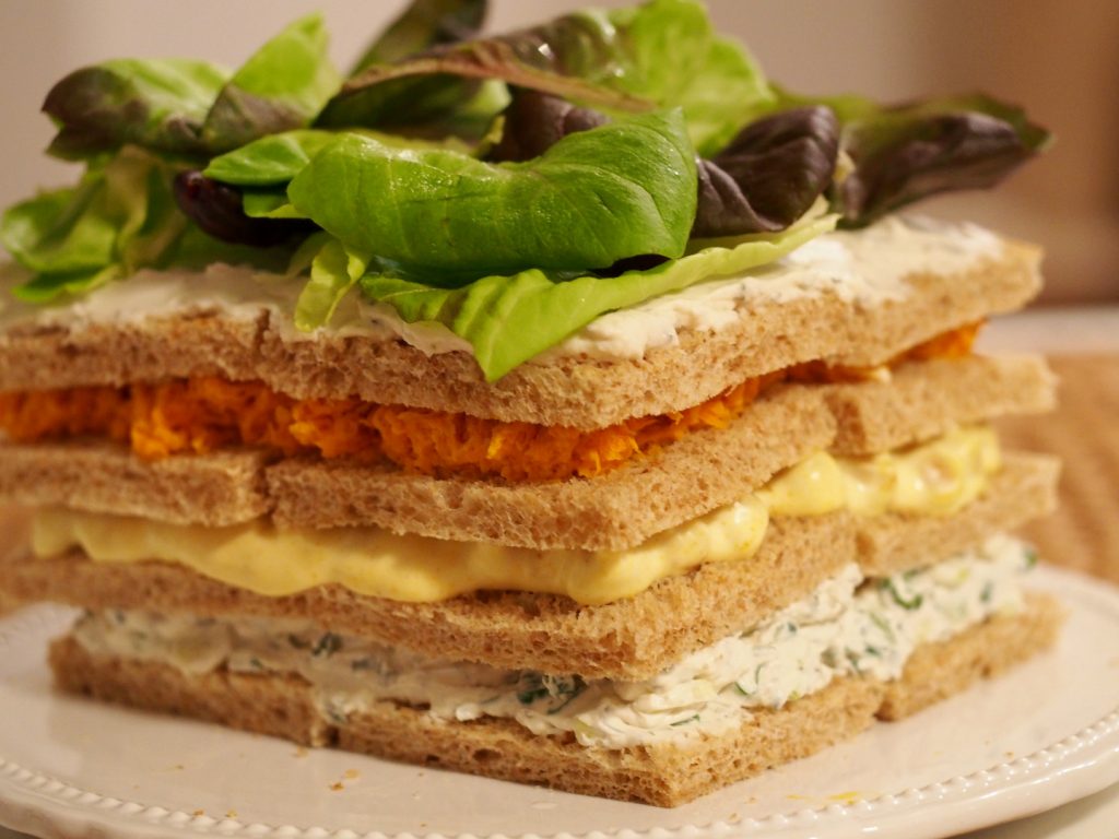 eine Sandwichtorte mit Gurkenbelag, Ei-Belag und Karottenbelag auf einer Tortenplatte. Die Sandwichtorte ist mit verschiedenen frischen Salatblättern dekoriert worden