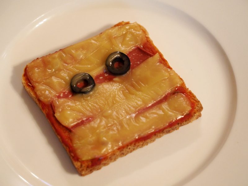Ein Toast mit veganer Salami, Käsestreifen und Olivenringe, gestaltet als Mumie