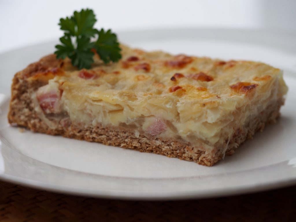 Ein Stück Zwiebelkuchen auf einem weißen Teller. Ein Zweiglein krause Petersilie liegt auf dem Zwiebelkuchenstück.