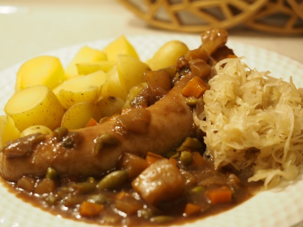 eine gekochte Weißwurst in Malz-Lebkuchensauce neben etwas Sauerkraut und einigen gekochten Kartoffelstücken