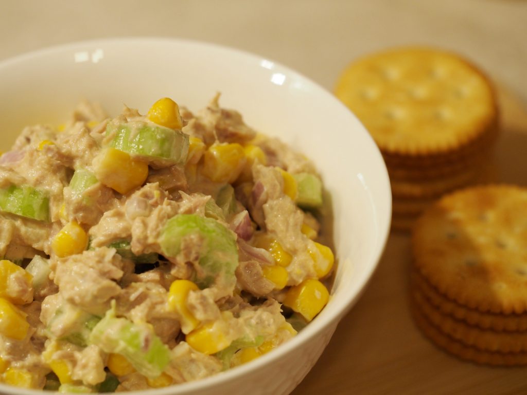 Eine kleine Schüssel mit dem Thunfischsalat mit Zwiebeln, Sellerie und Mais neben einigen aufgestapelten Crackern