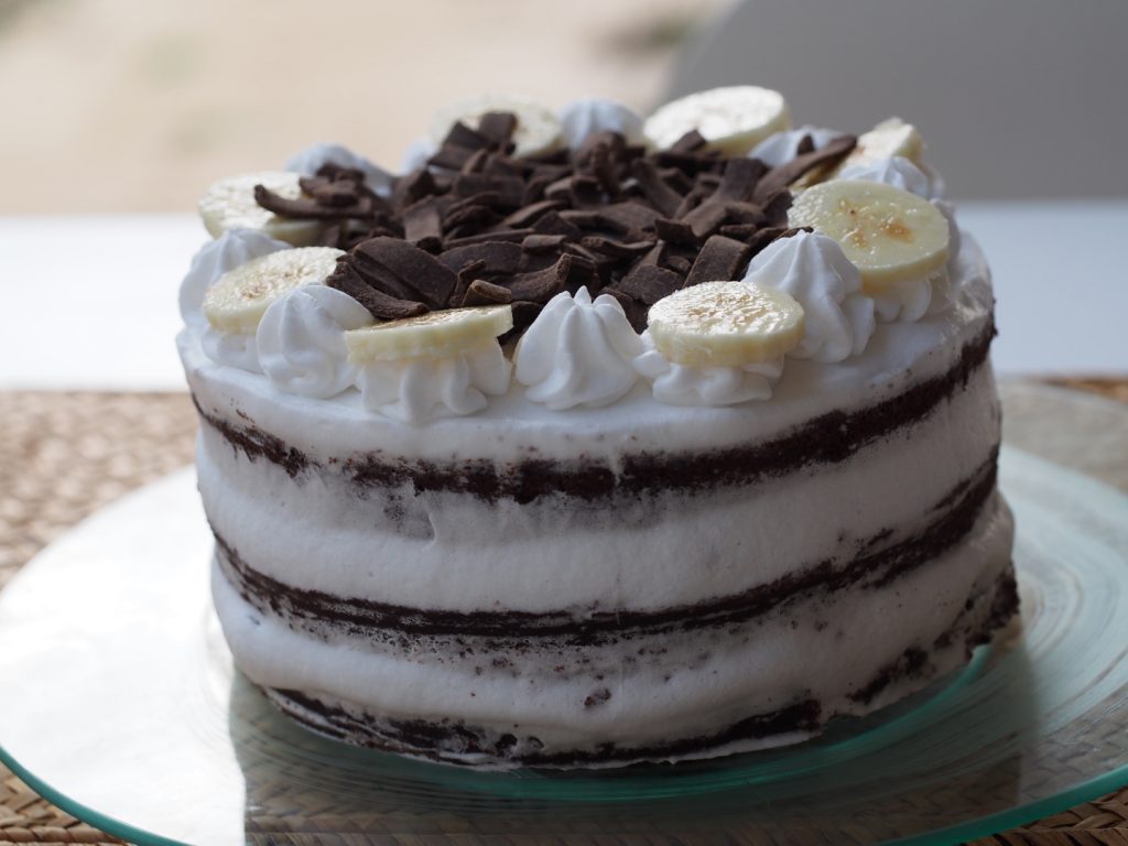 Die noch nicht angeschnittene Torte auf einer gläsernen Tortenplatte. Die Torte ist semi naked eingetrichen und oben mit Creme-Tuffs , Bananenscheiben und Schokosplitter dekoriert.