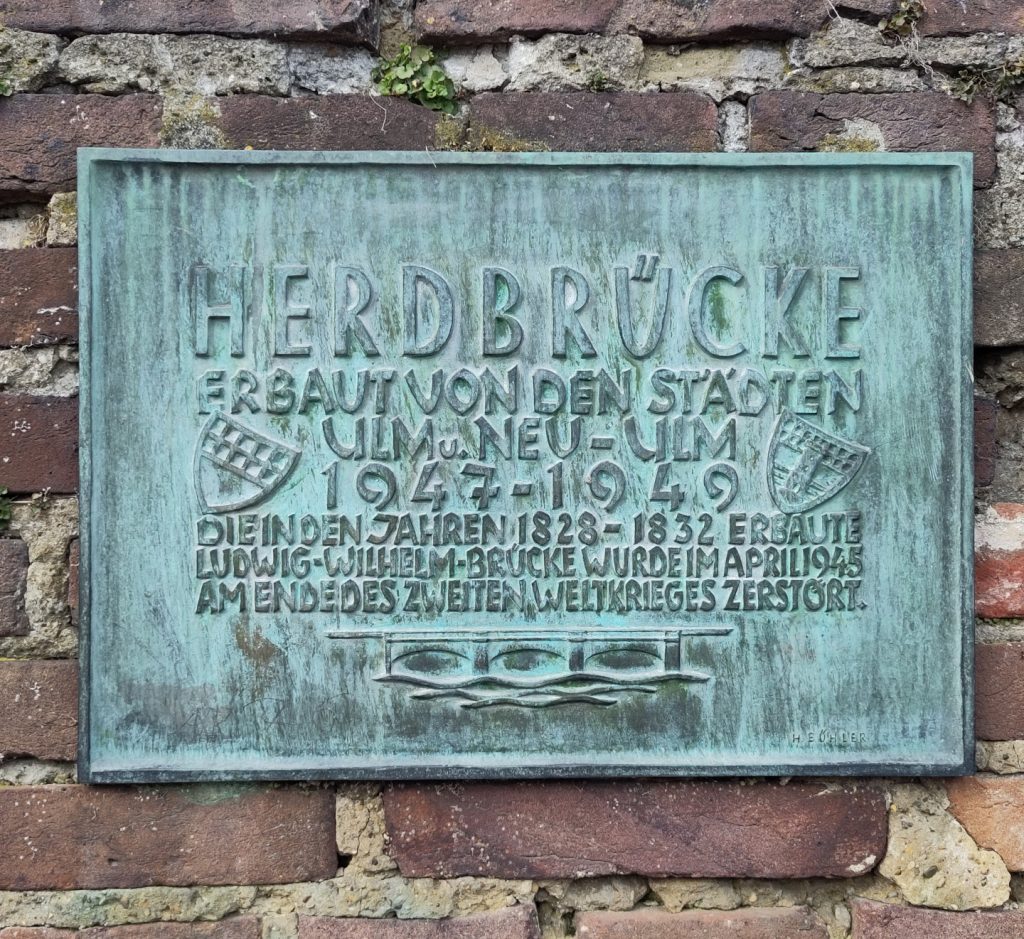 Ein Bronze Schild für die Herdbruckerstraße mit den Daten zum Bau:
Herdbrücke - erbaut von den Städten Ulm u. Neu-Ulm 1947-1949.
Die in den Jahren 1828-1832 erbaute Ludwig-Wilhelm-Brücke wurde im April 1945 am Ende des zweiten Weltkrieges zerstört.