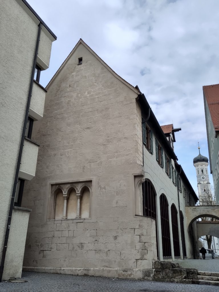 Rückseite des Steinhauses mit alten kirchlichen Zierelementen an einer ansonsten blanken Wand ohne Öffnungen.