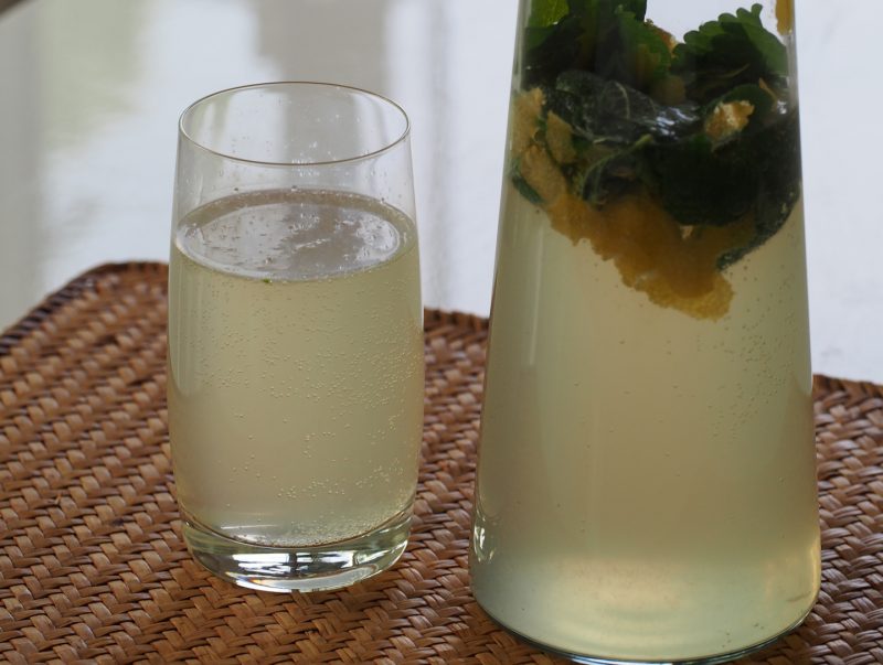 Eine halbvolle Karaffe neben einem Glas, beides gefüllt mit der Limonade, in der Karaffe schwimmen noch Zitronenmelissenblätter und Zitronenschalen.