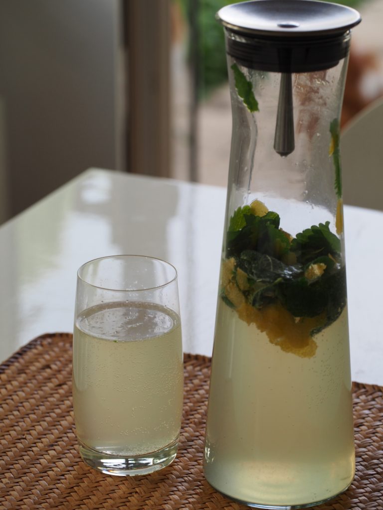 Eine halbvolle Karaffe neben einem Glas, beides gefüllt mit der Limonade, in der Karaffe schwimmen noch Zitronenmelissenblätter und Zitronenschalen.