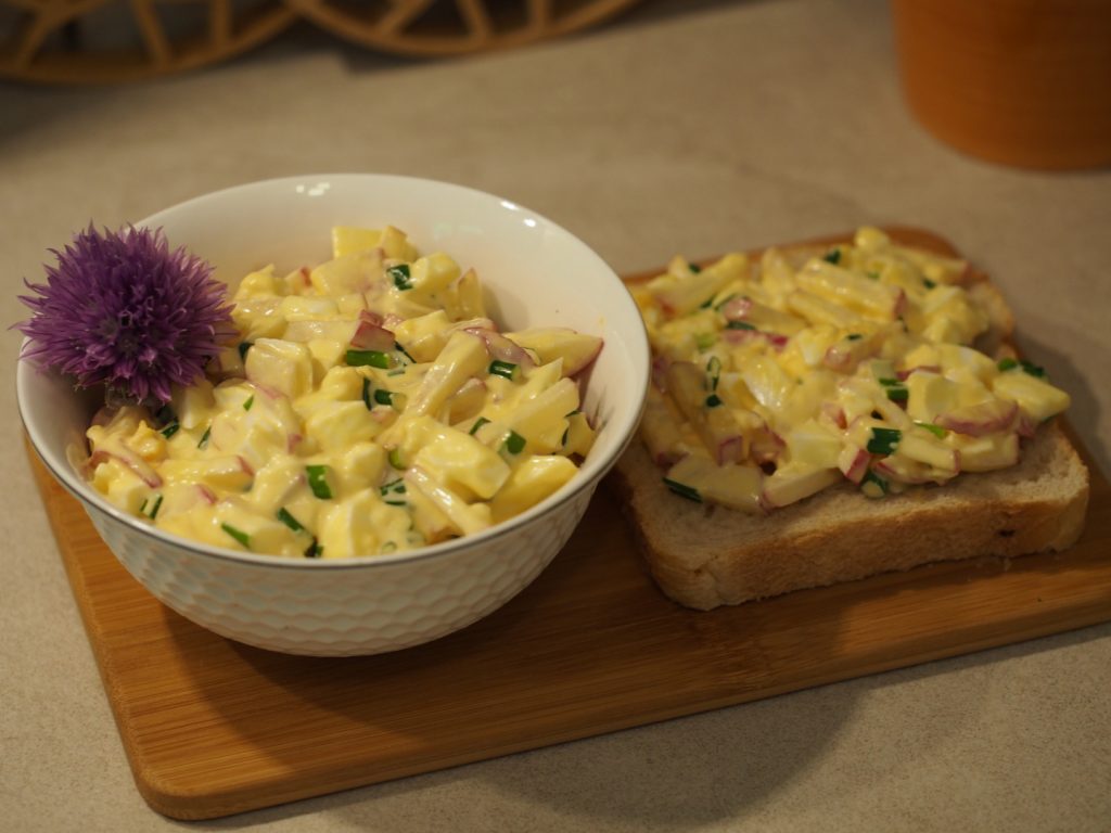 Die Schüssel mit dem Radieschen-Eiersalat auf einem Holzbrett neben einem mit dem Salat bestrichenem Brot.