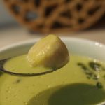 Eine grüne Spargelsuppe in einer Suppentasse, darüber wird ein Essläffel gehalten, auf dem eine Jakobsmuschel liegt.
