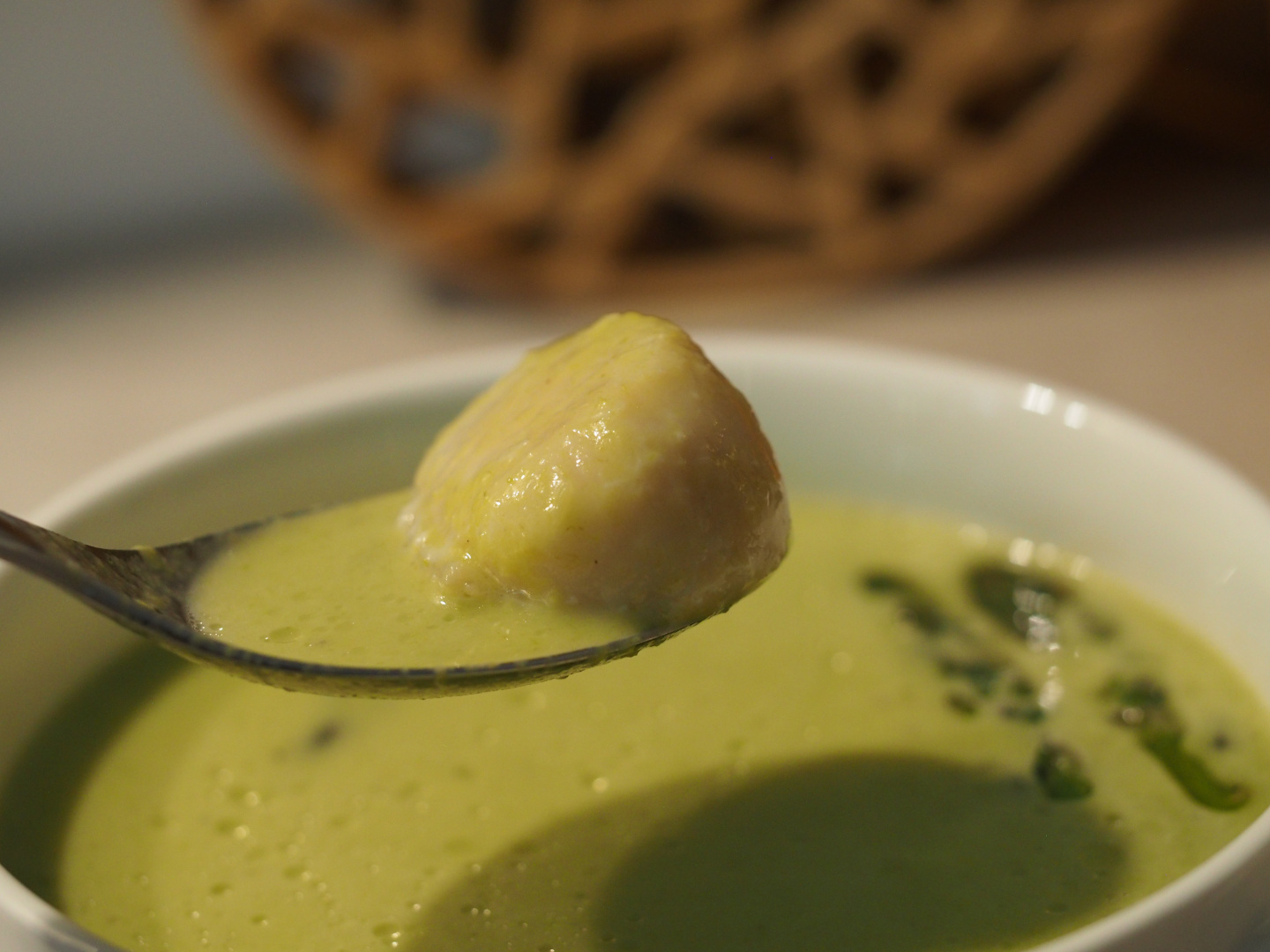 Eine grüne Spargelsuppe in einer Suppentasse, darüber wird ein Essläffel gehalten, auf dem eine Jakobsmuschel liegt.