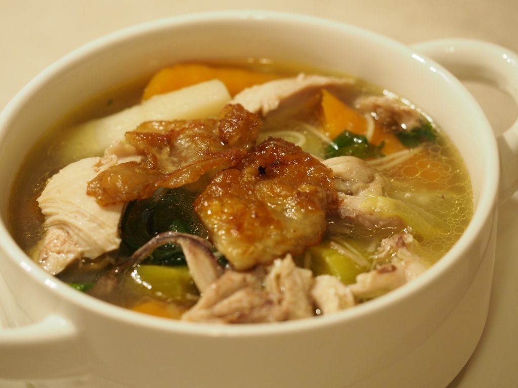 Eine Suppentasse gefüllt mit der Hühnersuppe mit Nudeln und einem Stück gebratener Hühnerhaut oben drauf