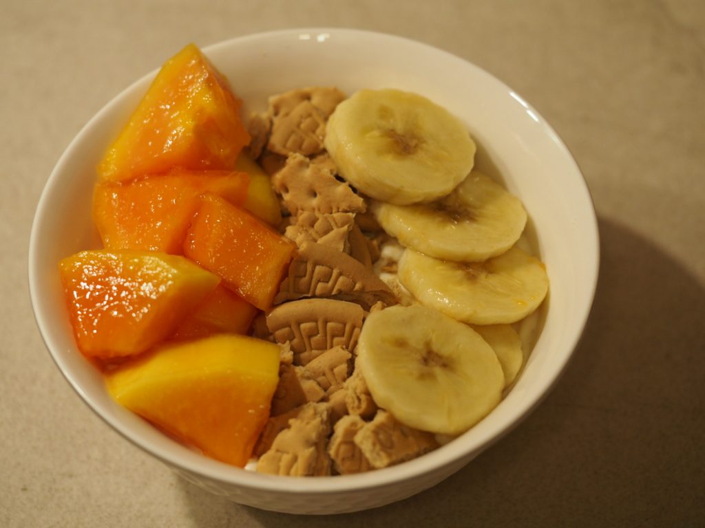 Eine kleine Schüssel mit dem Dessert. Auf der Oberfläche der Creme wurden Papayastücke und Bananenscheiben links und rechts einer Linie von Keksbruchstücken platziert.