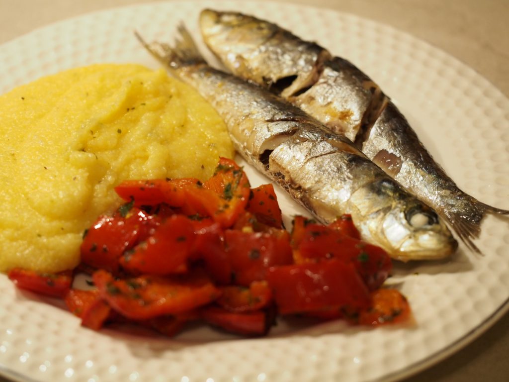 Zwei gebackene Sardinen auf einem Teller neben Polenta und Paprikagemüse
