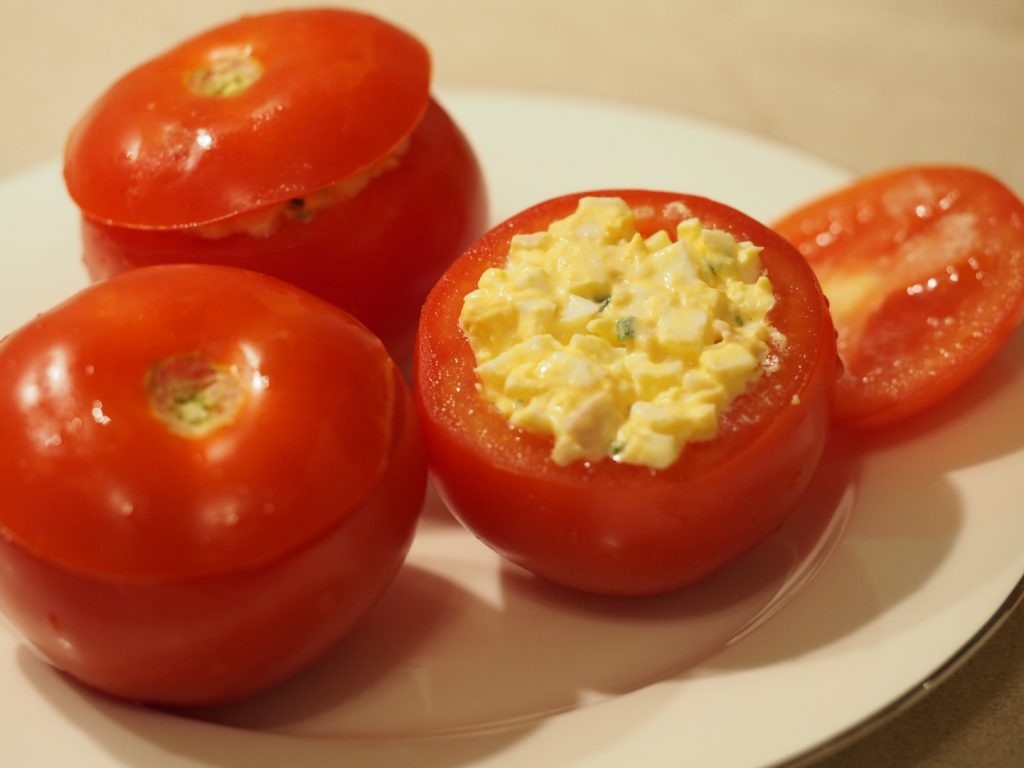 Drei gefüllte Tomaten auf einem Teller, dabei liegt der Deckel einer Tomate daneben, so dass die Füllung der Tomaten zu sehen ist.
