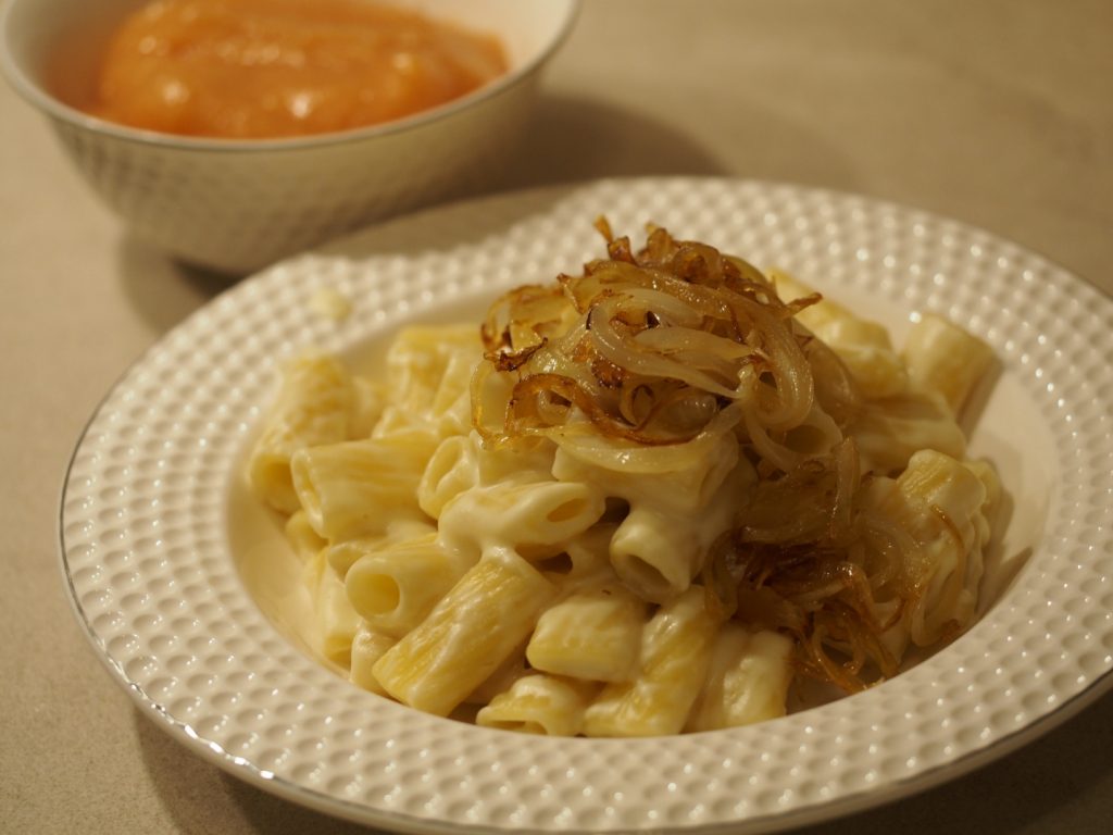Im Vordergrund ein Teller mit den Nudeln in Käsesauce mit gerösteten Zwiebeln darauf. Dahinter eine kleine Schüssel mit Quittenmus.