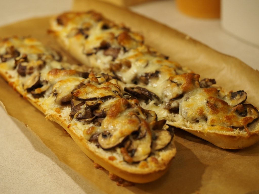 Zwei Baguette Hälften auf einem Stück Backpapier, die mit Champignons und Käse belegt und gebacken wurden.