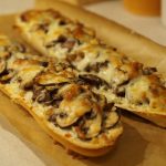 Zwei Baguette Hälften auf einem Stück Backpapier, die mit Champignons und Käse belegt und gebacken wurden.