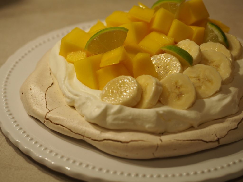 Die Pavlova auf einer weißen Tortenplatte. Die Pavlova ist mit aufgeschlagener Sahne bestrichen und diese wurde mit Mango-, Bananen- und Limettenstückchen garniert