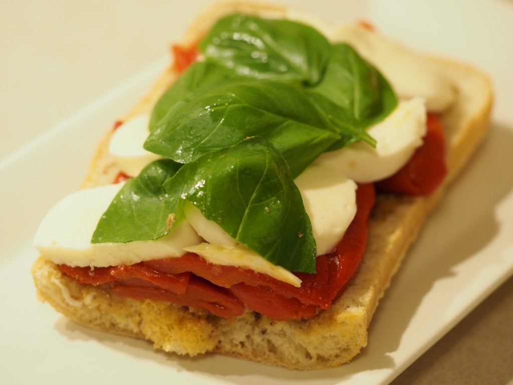 Das Sandwich ohne den oberen Brotdeckel gibt den Blick auf die frischen Basilikumblätter, den Mozzarella und die gerösteten Paprika frei