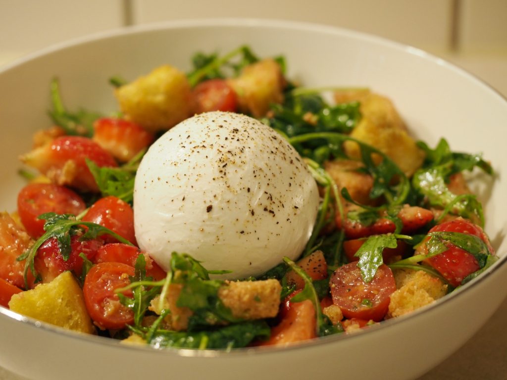 Eine große Portion des Salates in einer weißen Schüssel. In der Mitte liegt eine noch ganze Burrata Kugel, die mit gemahlenem Pfeffer bestreut wurde.
