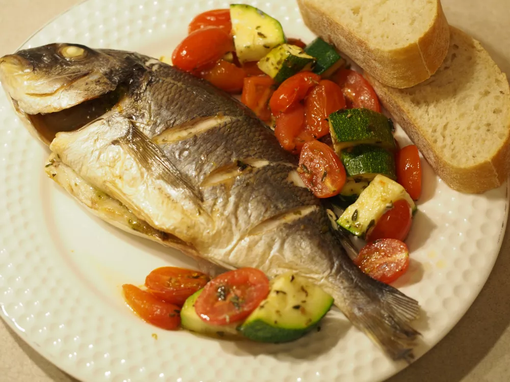 Eine gebackene ganze Dorade auf einem Teller. Daneben ist ein ebenso im Ofen gegartes Gemüse aus Tomaten und Zucchini, sowie zwei Scheiben eines Ciabatta.