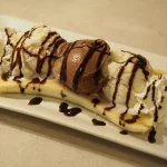 Ein selbst gemachter Bananen-Split mit eigenem Vanilleeis, Schokoladeneis, etwas Sprühsahne, Schokosauce und einer längshalbierten Banane auf einem rechteckigen weißen Teller. Für den Bananensplit wurden 2 Kugeln Vanilleeis und eine Kugel Schokoladeneis verwendet. Die Sahne ist jeweils am Ende der Bananen Eis-Reihe platziert.