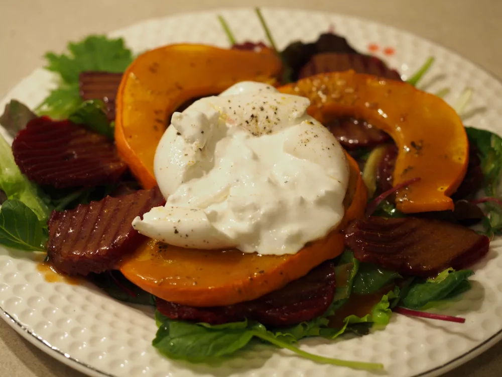 Ein Teller mit dem Salat. Zu unterst der grüne Salat, darauf die rote Bete und die gebackenen Kürbisspalten mit dem Dressing und in der Mitte liegt ein mit Pfeffer bestreuter Burrata. Der Burrata wurde nun auseinander gezogen, so dass das weiche Innere des Käses zu sehen ist.