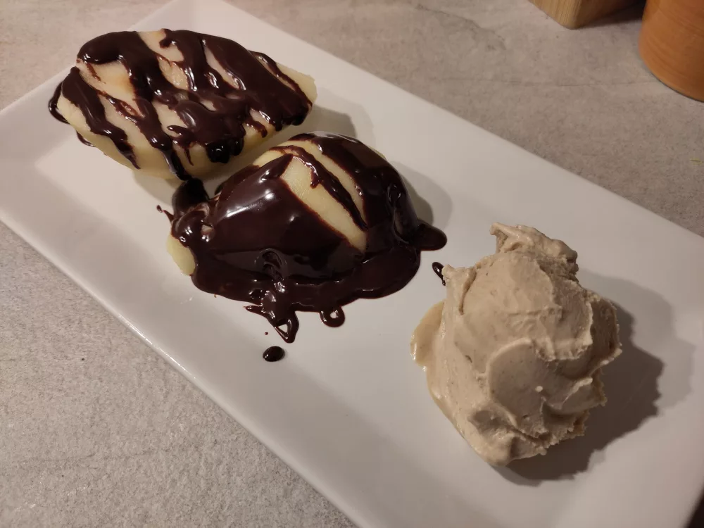 Zwei pochierte Birnenhälften auf einem rechteckigen weißen Teller, daneben eine Kugel Walnusseiscreme. Die Birnenhälften wurden großzügig mit Schokoladensauce übergossen.