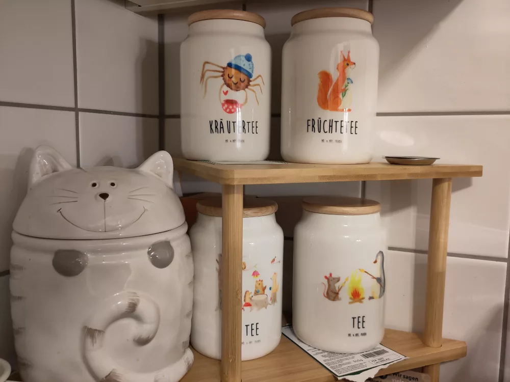 Auf einem kleinen Eckregal auf der Küchenarbeitsplatte stehen verschiedene Keramikdosen für Tee mit verschiedenen bunten Motiven. Unter manchen der Dosen liegt ein Zettel oder ein Abriss der ursprünglichen Teepackung. Daneben, auch noch auf dem Regal, steht auch eine Keramikdose in der Form einer Katze, in der das Trockenfutter für meine Kater gelagert wird.