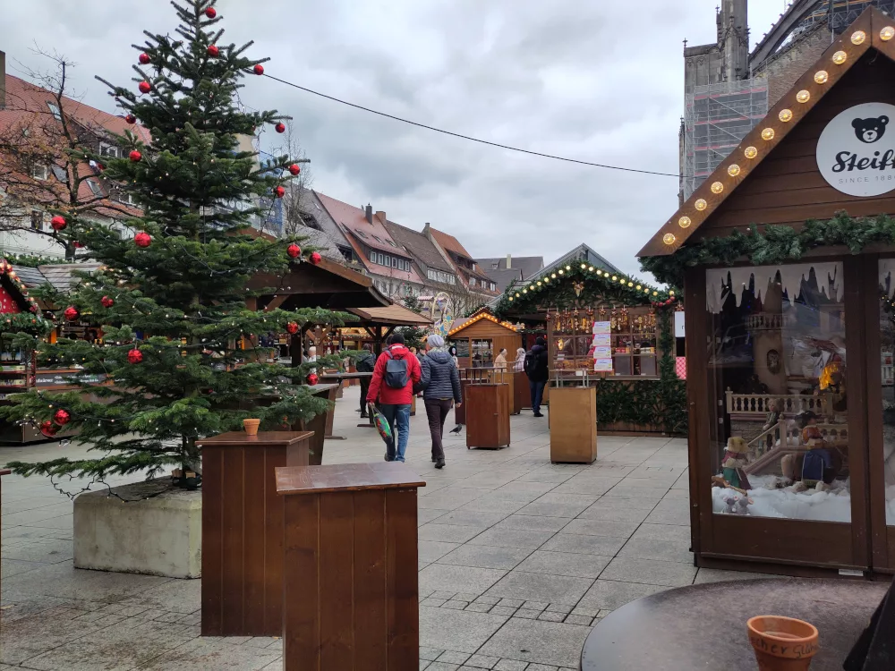Im Hintergrund ist ein kleines Riesenrad zu erkennen, davor verschiedene Stände und dekorierte Christbäume in Mitten von Stehtischen für die Gastronomie