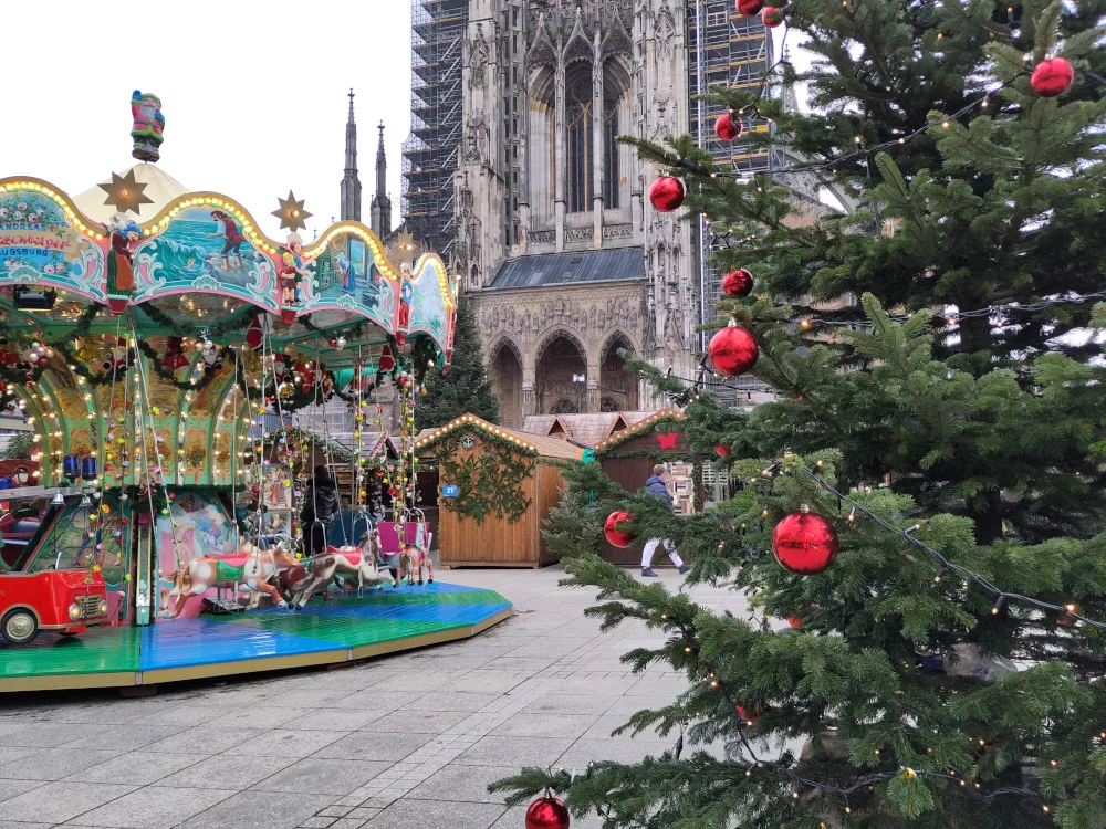 ein buntes Karussell mit Märchenmotiven auf dem Markt. Im Vordergrund ein Weihnachtsbaum, dekoriert mit roten Christbaumkugeln