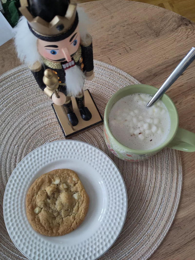 Ein heißer weißer Kakao mit Marshmallows aus so einer Schokoladenbombe, neben einem Teller auf dem ein Cookie liegt. Dekorativ wacht eine Nussknackerfigur über beides.