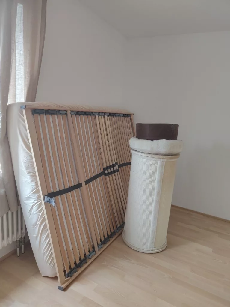 Im leeren Schlafzimmer lehnt die Matratze und der Lattenrost an der Wand, abgestützt durch eine Kratztonne der Kater, damit Matratze und Lattenrost nicht rutscht oder umkippt.