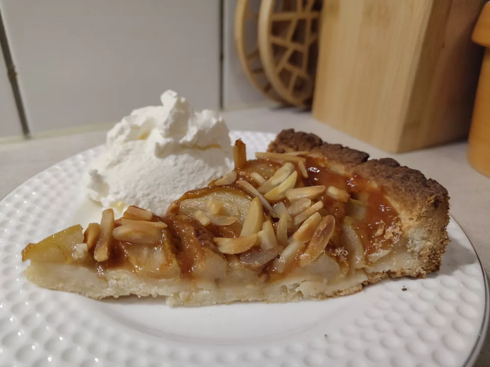 Ein Stück der Birnentarte von der Seite auf einem weißen Teller. Hinter dem Kuchenstück, wurde ein kleiner Berg Sahne platziert.