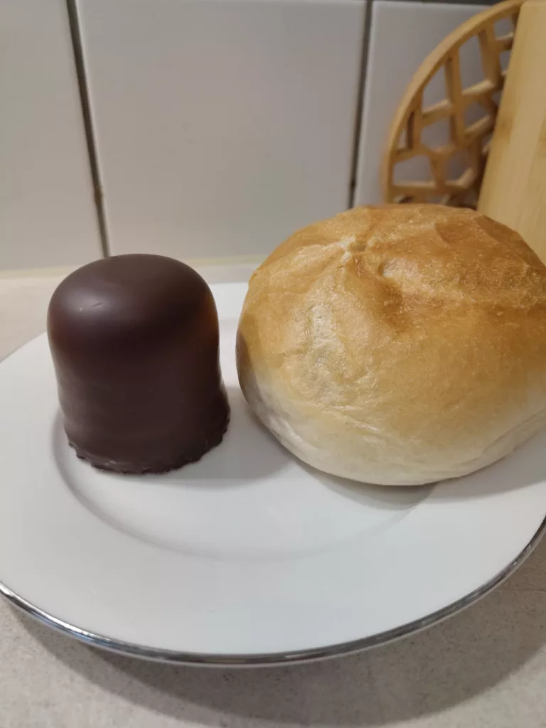 Dieser Schaumkuss, der gerade noch neben einer Semmel auf einem Teller steht, wird gleich zwischen den beiden Hälften der Semmel zerdrückt. Das Ergebnis ist ein Matschbrötchen oder eine Schokokuss-Semmel.