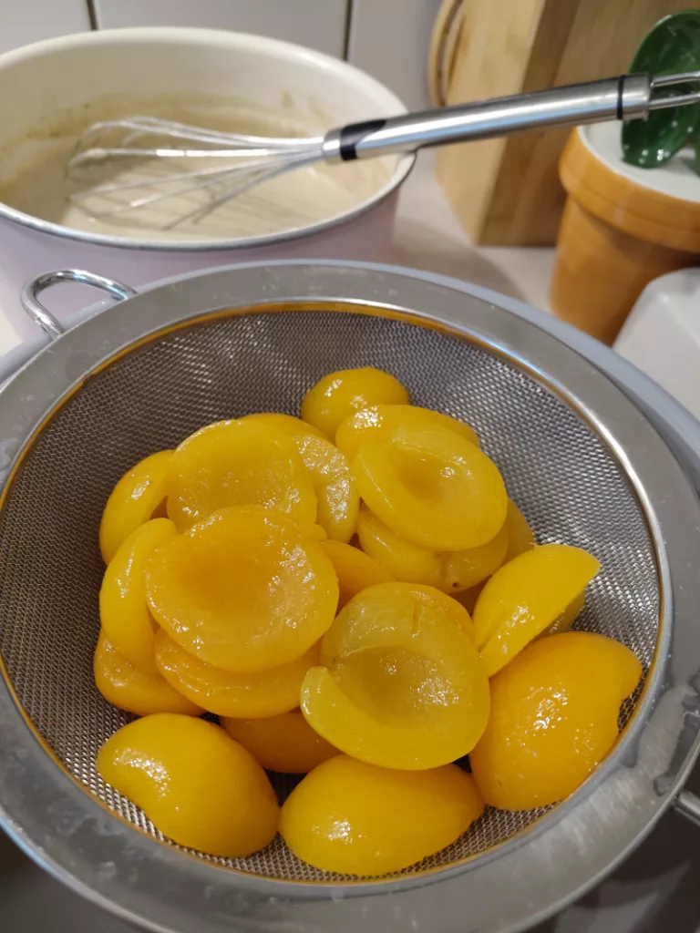 Hinten: Pudding in einem Kochtopf.
Vorne: eine Schüssel mit einem Sieb in dem Aprikosenhälften aus der Dose liegen zum Abtropfen.