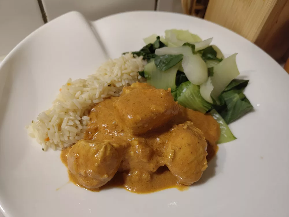 Eine Portion des Butter Chicken aus Pute mit einer orangenen Sauce neben etwas Reis und gedünstetem Pak Choi