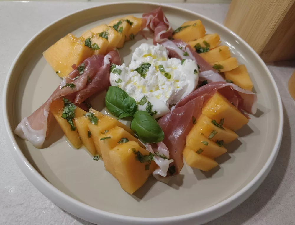 Ein Teller mit aufgeschnittener Cantaloupe-Melone in Stücken, dazwischen Prosciutto und in der Mitte ein geöffneter Burrata. Über alles wurde etwas von dem Dressing aus Basilikum, Limettensaft und Olivenöl gegeben. Ein Basilikumblattkranz wurde noch als Garnierung dazugelegt.
