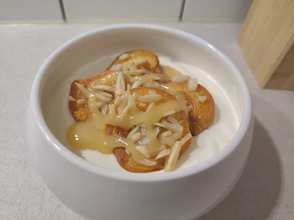 Eine Portion Joghurt in einer kleinen weißen Schüssel. Darauf wurden vier gebackene Aprikosenhälften mit Mandelsplitter gegeben. Darüber wurde ein cremiger Honig verteilt.